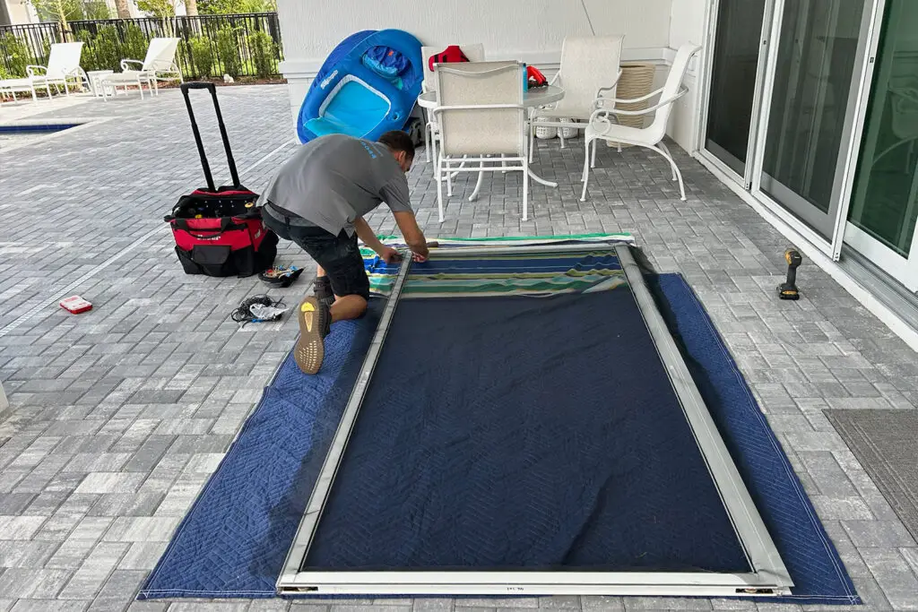 A person kneels on a blue padded mat, expertly handling tools from a red and black bag while assembling a large screen or frame. A blue pool float leans against the wall near the best sliding door repair setup. In the background, sliding glass doors open to an area paved with gray bricks.