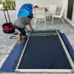 A person kneels on a blue padded mat, expertly handling tools from a red and black bag while assembling a large screen or frame. A blue pool float leans against the wall near the best sliding door repair setup. In the background, sliding glass doors open to an area paved with gray bricks.