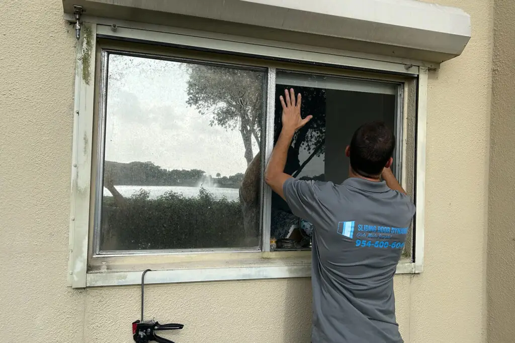 A person in a gray work shirt is adjusting a sliding window in a beige building, showcasing their expertise in patio door repair. Facing the window with trees and a lake visible through the glass, they have a toolkit on the windowsill. The shirt displays blue text and a phone number on the back.