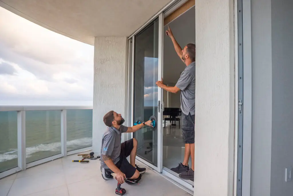 Two men are immersed in sliding door repair on a balcony overlooking the ocean. One squats, holding a tool, while the other points at the door frame. Scattered tools lie nearby as soft light filters through partly cloudy skies, highlighting their focused efforts in this picturesque setting.