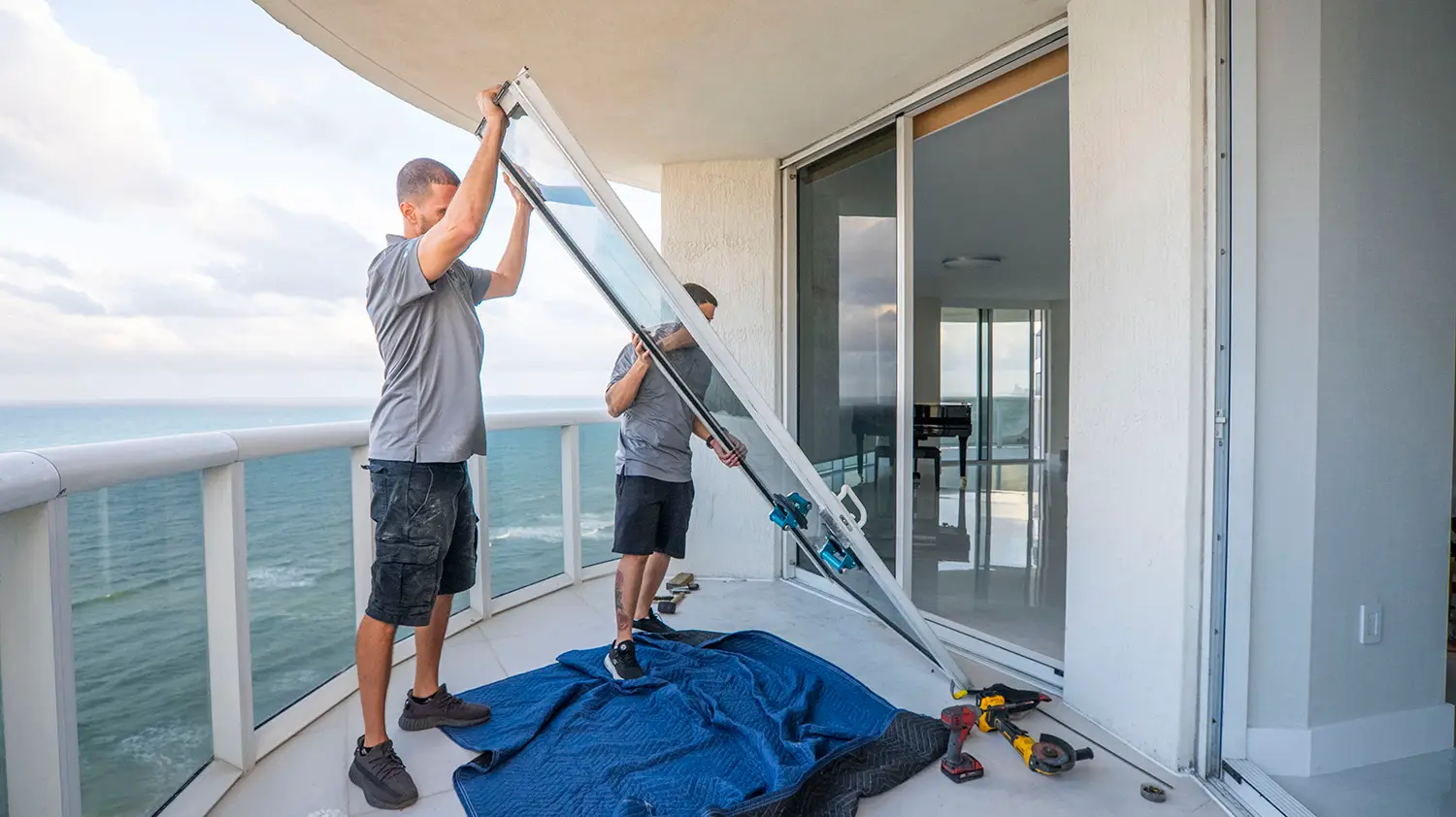 Two men expertly install a large window on a high-rise balcony overlooking the serene ocean. Dressed in gray shirts and shorts, they work efficiently, with a blue padded blanket cushioning the window frame amid tools for sliding door track repair. The sky is partly cloudy as the sea remains calm.