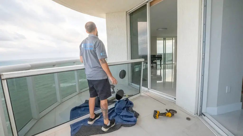 A man stands on a balcony with ocean views, holding a glass panel. He's wearing a gray shirt and black shorts. A drill and tools lie on the floor over a blue mat, poised for sliding door repair. Through the sliding glass door, indoor reflections reveal a piano. The sky is overcast.