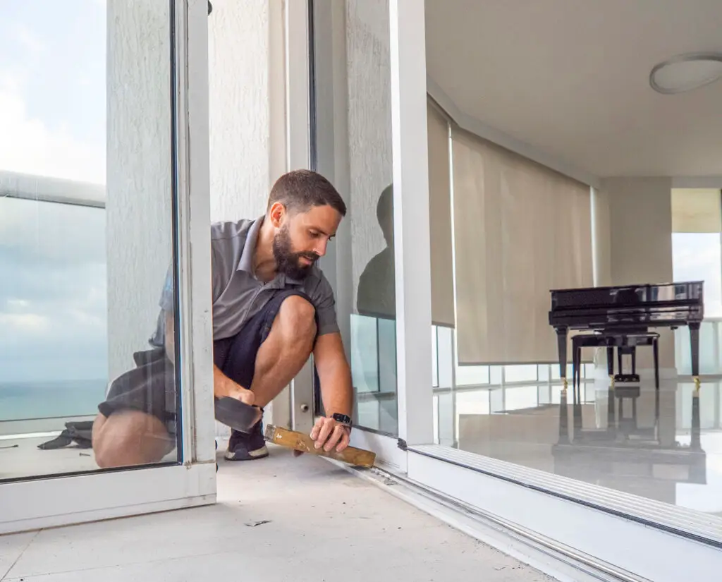 A bearded man in a gray shirt and black shorts kneels on one knee, skillfully performing sliding door repair. The bright room features a grand piano, while ocean views spill in through the windows.