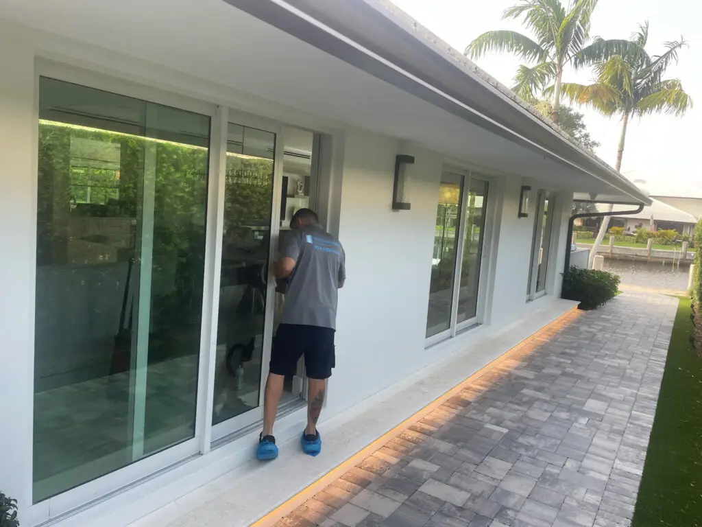 A man wearing a gray shirt and blue shoes peers through a large window of a modern white house. The house, needing sliding door repair, features multiple glass doors and a gray brick path. Lush greenery, including palm trees, borders the scene. A strip of light runs along the house’s base.