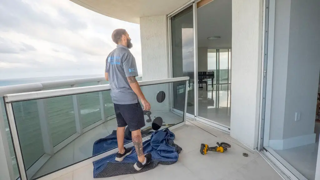 A man with tattoos stands on a high-rise balcony, holding a black disc. He's wearing a blue shirt and black shorts. A power tool, possibly for sliding door repair, is on the ground beside him, and a blue pad covers part of the floor. The ocean is visible in the background through glass walls.