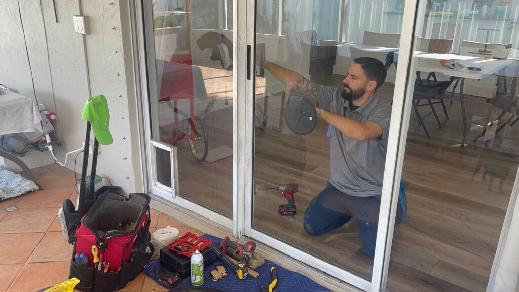 A man kneels, focused on sliding door track repair, using a power drill amid tools like a toolbox and screwdriver. Indoors, with a chair and dog visible through the glass patio door, he works diligently. A towel covers the floor to catch debris as he sprays cleaning solution.