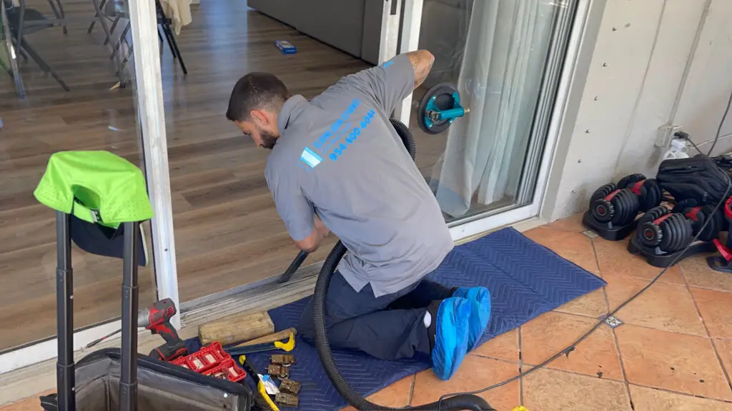 A man in a gray shirt and blue shoe covers is kneeling on a blue mat, expertly focused on sliding door repair with a vacuum. Nearby, weights, a toolbox, and various cleaning tools lie within reach as sunlight from the window illuminates his precise work.