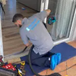 A man in a gray shirt and blue shoe covers is kneeling on a blue mat, expertly focused on sliding door repair with a vacuum. Nearby, weights, a toolbox, and various cleaning tools lie within reach as sunlight from the window illuminates his precise work.