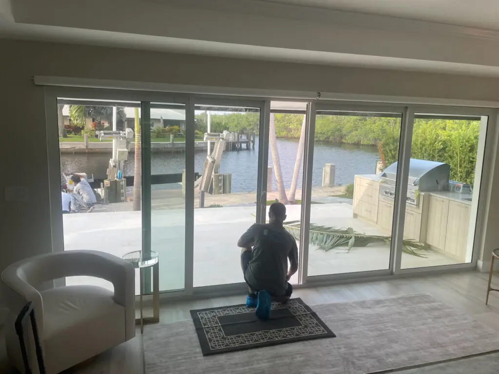 Crouching indoors near the sliding glass doors, a person gazes out at a waterfront view featuring a dock and canal. Outside, another sits by the water. A grill and fallen palm frond dot the patio. The room's elegance is enhanced by a white armchair and patterned rug, all framed by flawless sliding door repair.