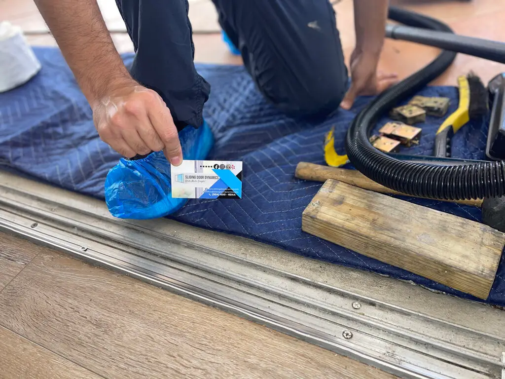 A person kneels on a blue padded mat, placing a card next to their knee, preparing for sliding door track repair. They wear a blue shoe cover and dark pants. Nearby are tools, including a vacuum hose, wooden blocks, and a yellow utility knife—all set for indoor flooring work near the metal track.