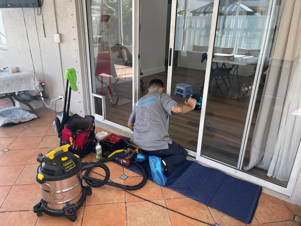 A technician in a gray shirt and blue shoe covers is kneeling on a mat by a sliding glass door, diligently performing sliding door repair. He uses a drill on the door's frame, surrounded by tools, a red toolbox, and a vacuum cleaner. Inside, wooden flooring complements the blue stool; outside, terracotta tiles shine.