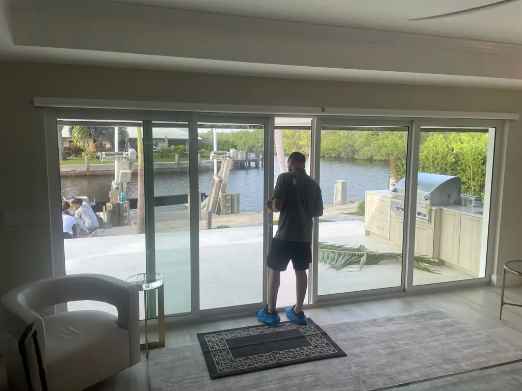 A person stands in front of large sliding glass doors, gazing out onto a patio and waterway with docks. Wearing a gray shirt, black shorts, and blue shoes, they contemplate the scene. Inside are a white chair and rug; outside, plants flourish next to the barbecue grill awaiting any necessary sliding door track repair.