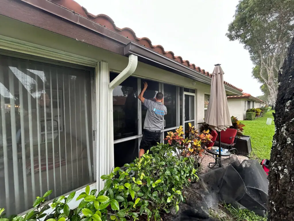 A person is installing mesh screens on the windows of a house with a tiled roof while surrounded by lush plants, including shrubs and a tree. Nearby, patio furniture and a large umbrella complement the scene. In the distance, sliding door repair work takes place as clouds blanket the sky.