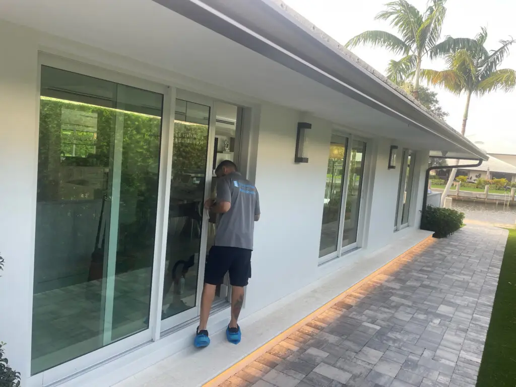 A man in a gray shirt and black shorts, wearing blue shoe covers, carefully tends to the patio door repair of a modern white house. The pathway is paved with gray bricks, and palm trees are visible in the background. Warm lighting highlights the home's sleek design.