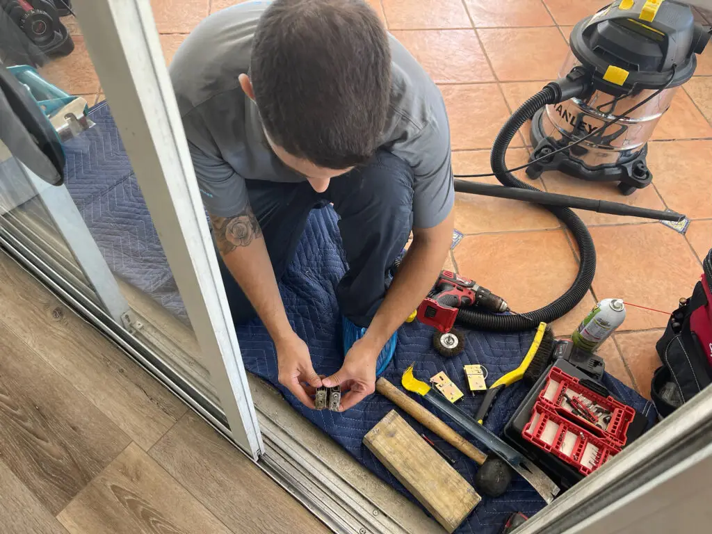 Kneeling indoors by an open door, a person with short brown hair is focused on the best sliding door repair solutions. Tools, a vacuum cleaner, and a red toolbox are scattered on the blue mat below them, set against a blend of wood and tiled surfaces.