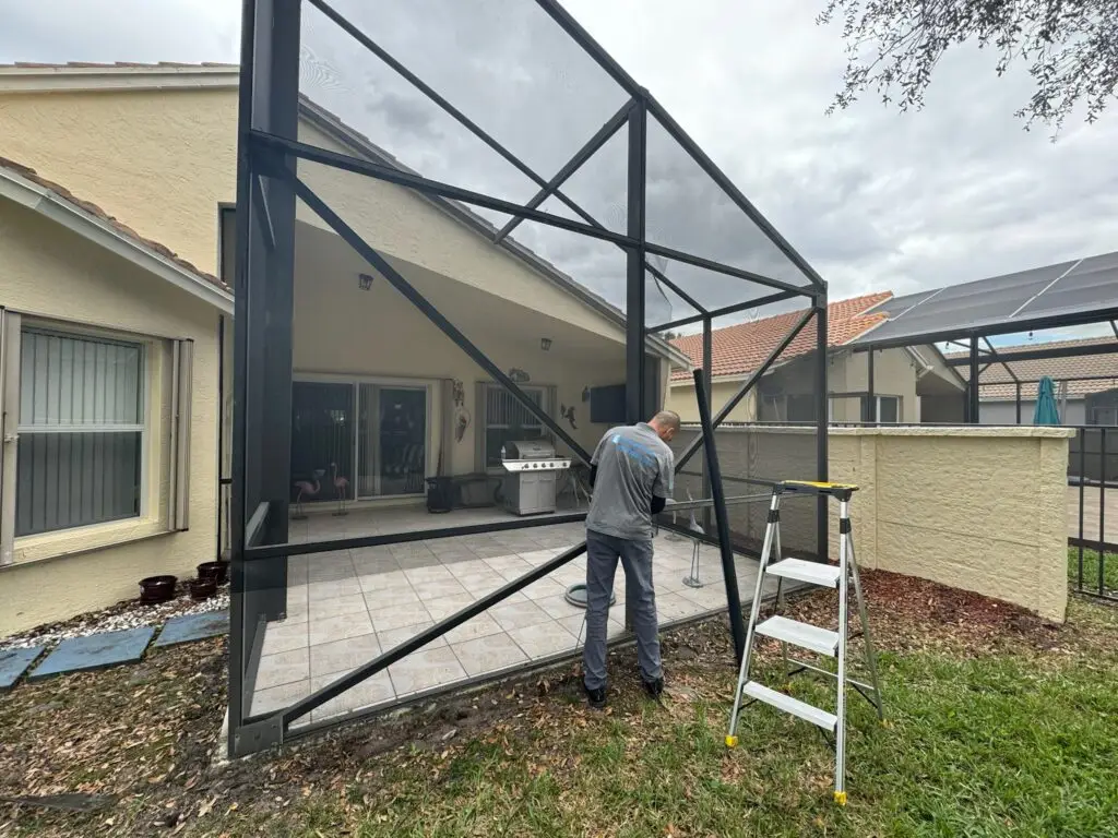 A person is repairing a large, metal-framed screen by the sliding door on a back patio of a beige house. Nearby, a foldable ladder rests on the grass. The patio has tiled flooring with outdoor furniture and a grill. Solar panels adorn the neighboring roof under a cloudy sky.