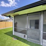 A screened-in patio attached to a house features a mesh door and cat door. The patio overlooks a grassy backyard with a wooden privacy fence. A blue sky with clouds is visible. A few construction materials and gloves hint at some patio door repair work in progress on the floor.