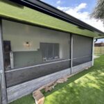 A worker is installing a screen on a porch with large glass sliding doors, highlighting the need for occasional sliding door repair. The porch has a wall-mounted light and partly painted green wall, while the floor is protected with paper. Outside, there's artificial grass and a wooden fence beneath partly cloudy skies.