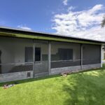 A single-story house with a screened porch faces a lawn covered in artificial grass. The sky is partly cloudy with patches of blue. Tools and building materials are scattered near the porch, suggesting ongoing sliding door track repair. A wooden fence surrounds the yard.