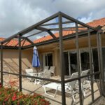 a patio with a metal frame structure under construction, featuring a glass patio door leading into a house with a terracotta roof. white patio furniture, including chairs and a table with a blue umbrella, is arranged on the paved area. red flowering bushes border the south florida screen enclosure.