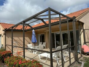 a patio with a metal frame structure under construction, featuring a glass patio door leading into a house with a terracotta roof. white patio furniture, including chairs and a table with a blue umbrella, is arranged on the paved area. red flowering bushes border the south florida screen enclosure.