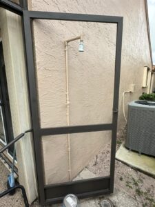 a metal frame door leans against a textured beige wall outdoors under a south florida screen enclosure. behind it, a showerhead is mounted on vertical pipes attached to the wall. an air conditioning unit is visible to the right, and a potted plant peeks out on the left over concrete ground scattered with debris.