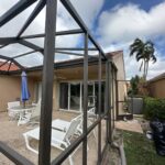 a patio area featuring white metal outdoor furniture, including chairs and a table, set on a pebble textured surface. a blue umbrella and a south florida screen enclosure surround the area. a beige house with sliding glass doors, a palm tree, and a partly cloudy sky complete the serene view.