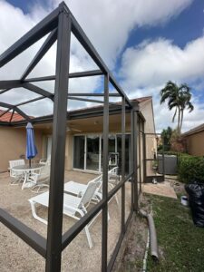 a patio area featuring white metal outdoor furniture, including chairs and a table, set on a pebble textured surface. a blue umbrella and a south florida screen enclosure surround the area. a beige house with sliding glass doors, a palm tree, and a partly cloudy sky complete the serene view.