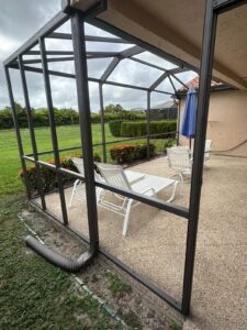 patio enclosed with a dark metal frame and screened panels near a beige house. it contains white loungers, a small table, and a blue closed umbrella. outside, there's green lawn and neatly trimmed hedges under a cloudy sky. a downspout is visible at the corner, suggesting recent screen enclosure repair.