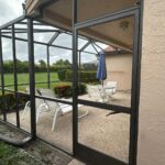 a patio under a screen enclosure restoration, enclosed with a black metal frame and screens. inside, white lounge chairs and an upright blue umbrella surround a small table. the patio is embraced by grass and hedges. an overcast sky looms above, visible through the screens and landscaped area.