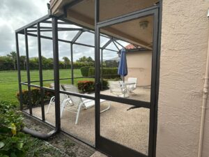 a patio under a screen enclosure restoration, enclosed with a black metal frame and screens. inside, white lounge chairs and an upright blue umbrella surround a small table. the patio is embraced by grass and hedges. an overcast sky looms above, visible through the screens and landscaped area.