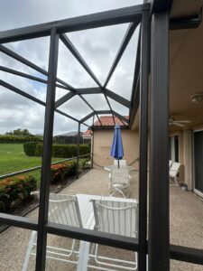 a screened patio with two white lounge chairs and a white table is shown, embodying the essence of a south florida screen enclosure. an umbrella shaded in blue stands beside them, while greenery and bushes offer a lush backdrop. the house features a tan exterior and red tiled roof under cloudy skies.