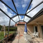 a patio beneath a metal frame awaits its screen enclosure repair. it features white furniture, a table with a blue umbrella, and a ceiling fan. a ladder stands on the grass under partly cloudy skies. bushes and trees surround the lawn, while the house boasts a red tiled roof and sliding glass doors.