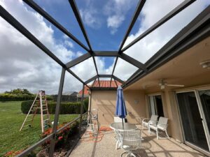 a patio beneath a metal frame awaits its screen enclosure repair. it features white furniture, a table with a blue umbrella, and a ceiling fan. a ladder stands on the grass under partly cloudy skies. bushes and trees surround the lawn, while the house boasts a red tiled roof and sliding glass doors.