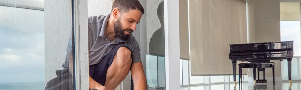 A bearded man in a gray shirt and black shorts kneels on one knee, skillfully performing sliding door repair. The bright room features a grand piano, while ocean views spill in through the windows.