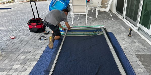 A person kneels on a blue padded mat, expertly handling tools from a red and black bag while assembling a large screen or frame. A blue pool float leans against the wall near the best sliding door repair setup. In the background, sliding glass doors open to an area paved with gray bricks.