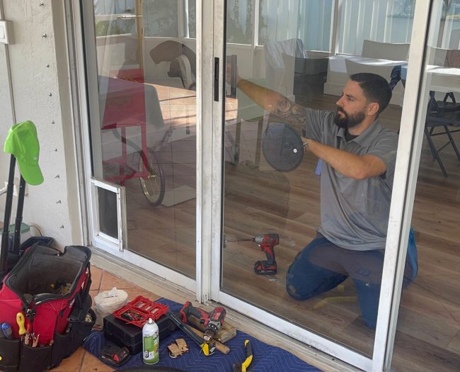 A man kneels, focused on sliding door track repair, using a power drill amid tools like a toolbox and screwdriver. Indoors, with a chair and dog visible through the glass patio door, he works diligently. A towel covers the floor to catch debris as he sprays cleaning solution.