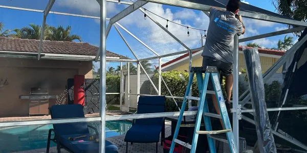 A man stands on a blue ladder, focusing on patio door repair within a screened enclosure around a backyard pool area. Patio furniture, including two lounge chairs and a small table, are inside the space. A partially cloudy sky and trees create a serene backdrop.