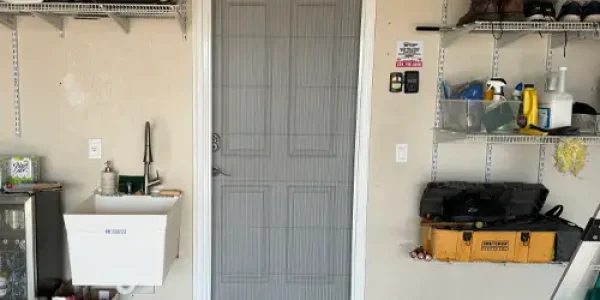 A closed door with a mesh screen, framed by a white border, leads into the garage. Shelves on the right hold boots, tools, and cleaning supplies. On the left are a sink and a small cabinet. A clipboard and “No Parking” sign hang next to the door that might need some sliding door track repair soon.