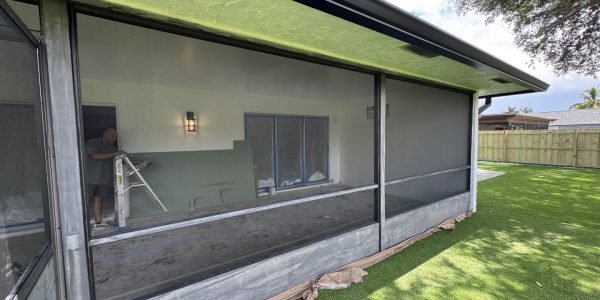 A person on a ladder works on the interior of a screened porch with a green ceiling, ensuring smooth operation through sliding door track repair. The porch has large mesh screens and is part of a house with sliding glass doors. The floor is partially covered to protect it against debris, while artificial grass and a wooden fence frame the yard outside.