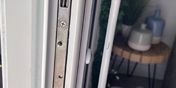 Close-up of a white door showcasing a secure metal lock mechanism and matching handle. In the background, a wooden table with potted plants and a fan on the floor suggests an indoor setting bathed in natural light.