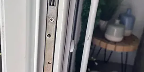 Close-up of a white door showcasing a secure metal lock mechanism and matching handle. In the background, a wooden table with potted plants and a fan on the floor suggests an indoor setting bathed in natural light.