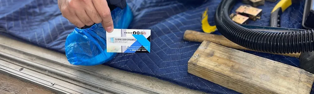 A person kneels on a blue padded mat, placing a card next to their knee, preparing for sliding door track repair. They wear a blue shoe cover and dark pants. Nearby are tools, including a vacuum hose, wooden blocks, and a yellow utility knife—all set for indoor flooring work near the metal track.