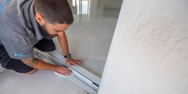 A man with a beard is expertly installing a sliding glass door. Kneeling on the tiled floor in his gray shirt and dark shorts, he's focused on sliding door track repair. His left hand adjusts the frame while his right holds a tool. A wristwatch glints on his wrist against the white wall backdrop.